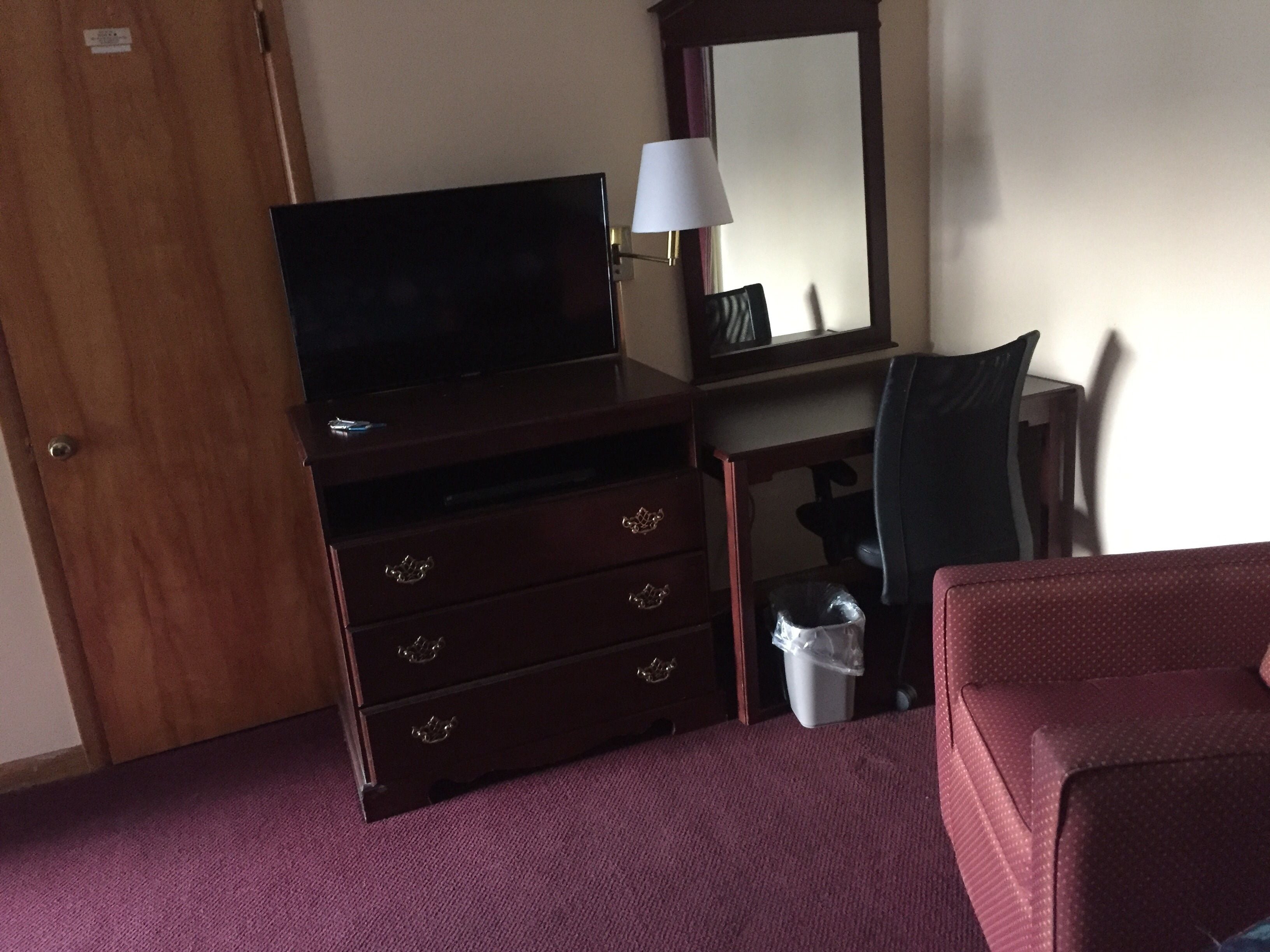 A hotel room with a wooden dresser, a television, a desk with a chair, a lamp, and a trash bin.