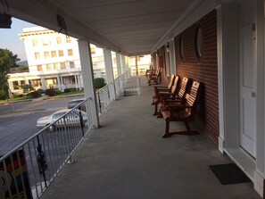 A covered porch with wooden benches, a metal railing, and a view of a street with parked cars. - Motel Townhouse (Bedford)