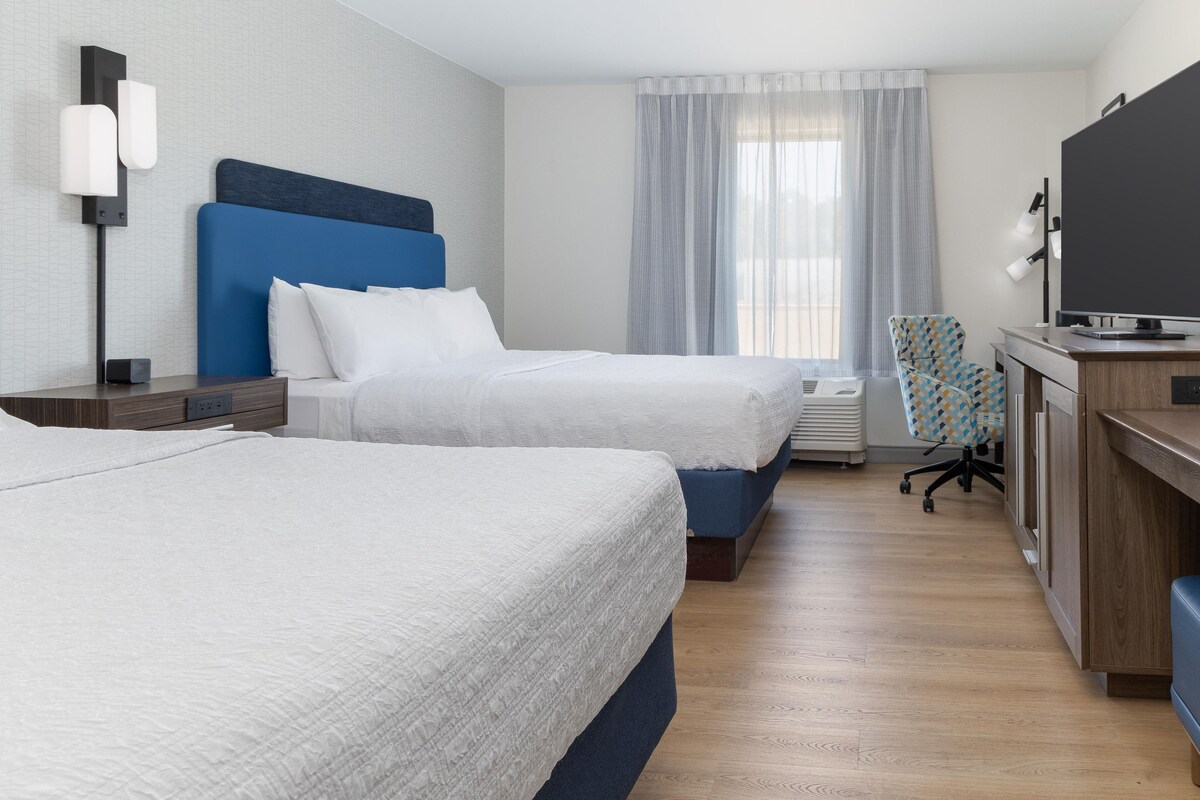 Una habitació d'hotel amb dos llits, un escriptori amb televisió de pantalla plana, una cadira i una finestra amb cortines.