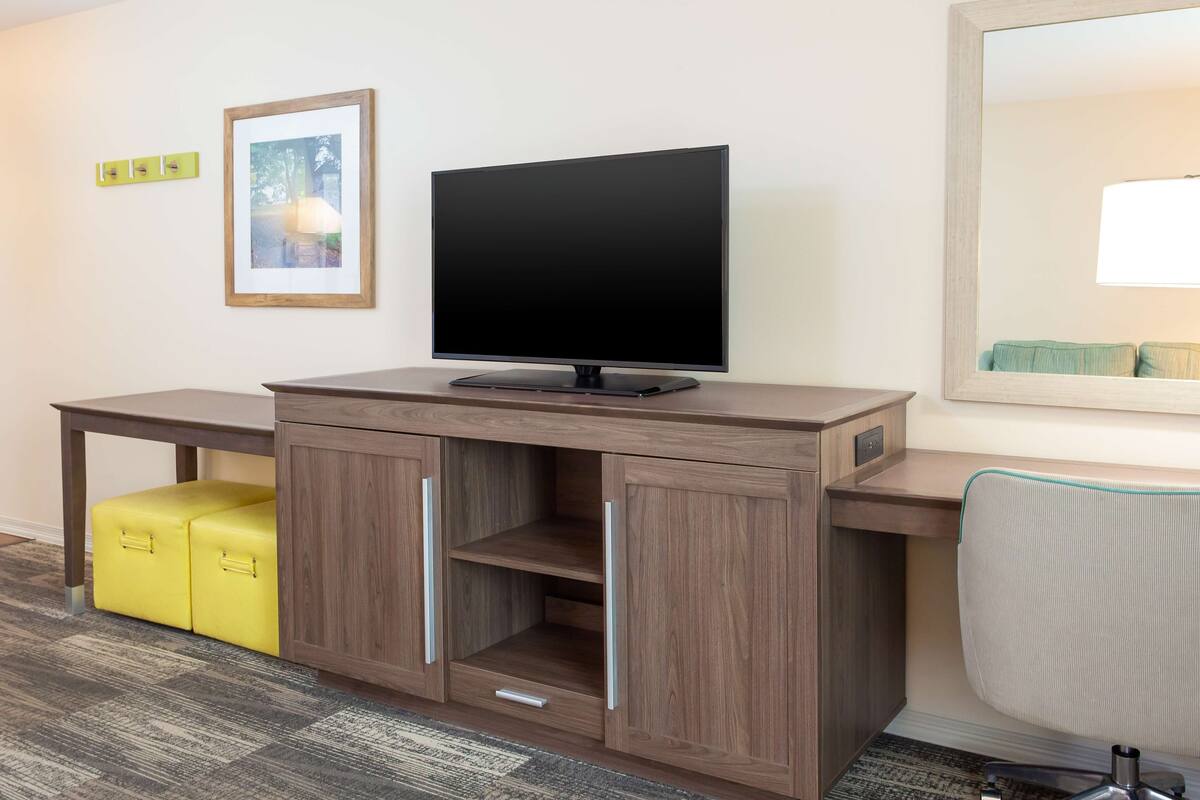 Quarto moderno com escrivaninha de madeira, televisão de tela plana, um banquinho amarelo e um espelho grande.