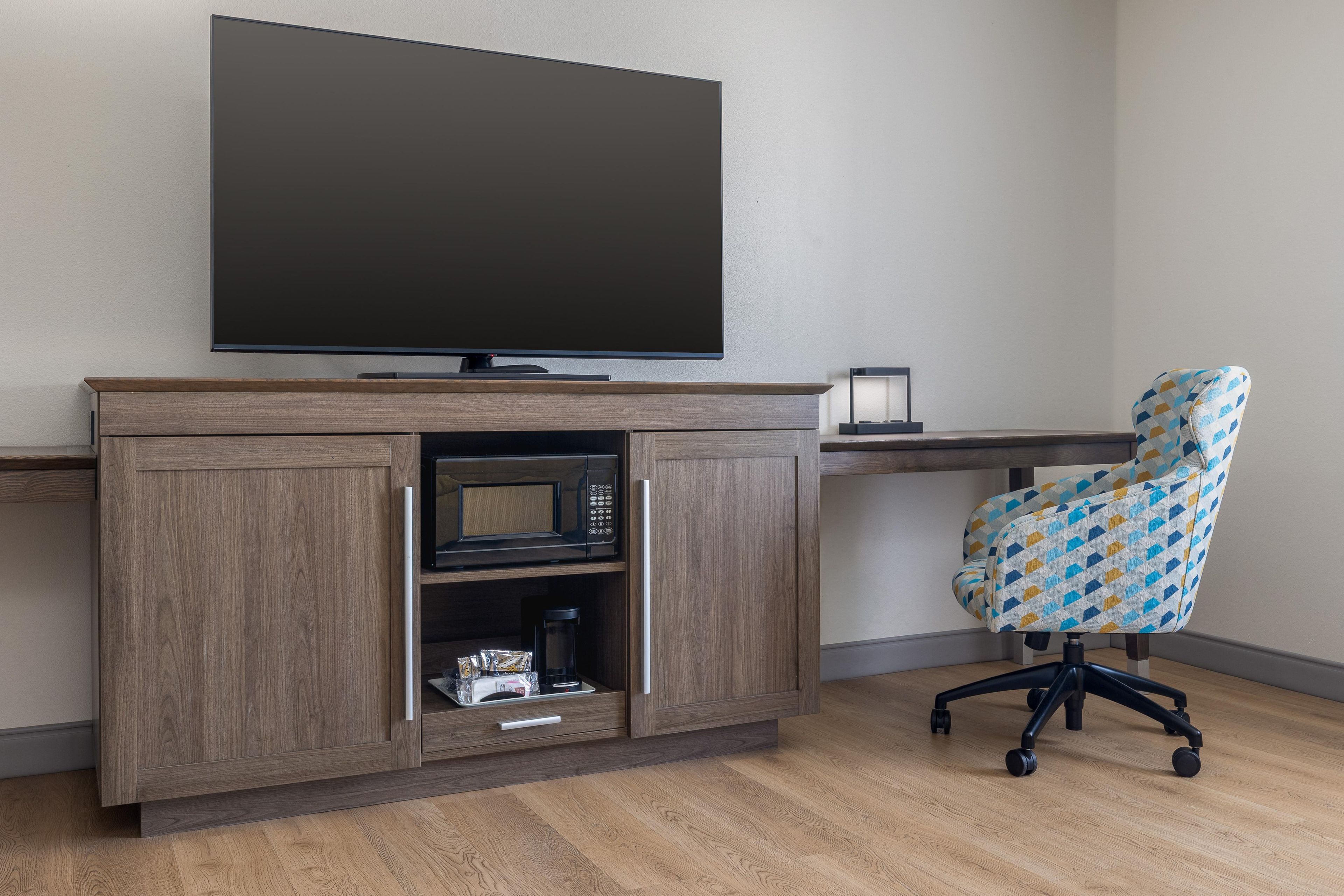 A modern room with a flat-screen TV mounted on the wall, a wooden desk with a built-in microwave, and a patterned office chair.