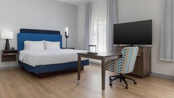 Habitación de hotel con cama, escritorio, silla, televisor, lámpara y ventana con cortinas.