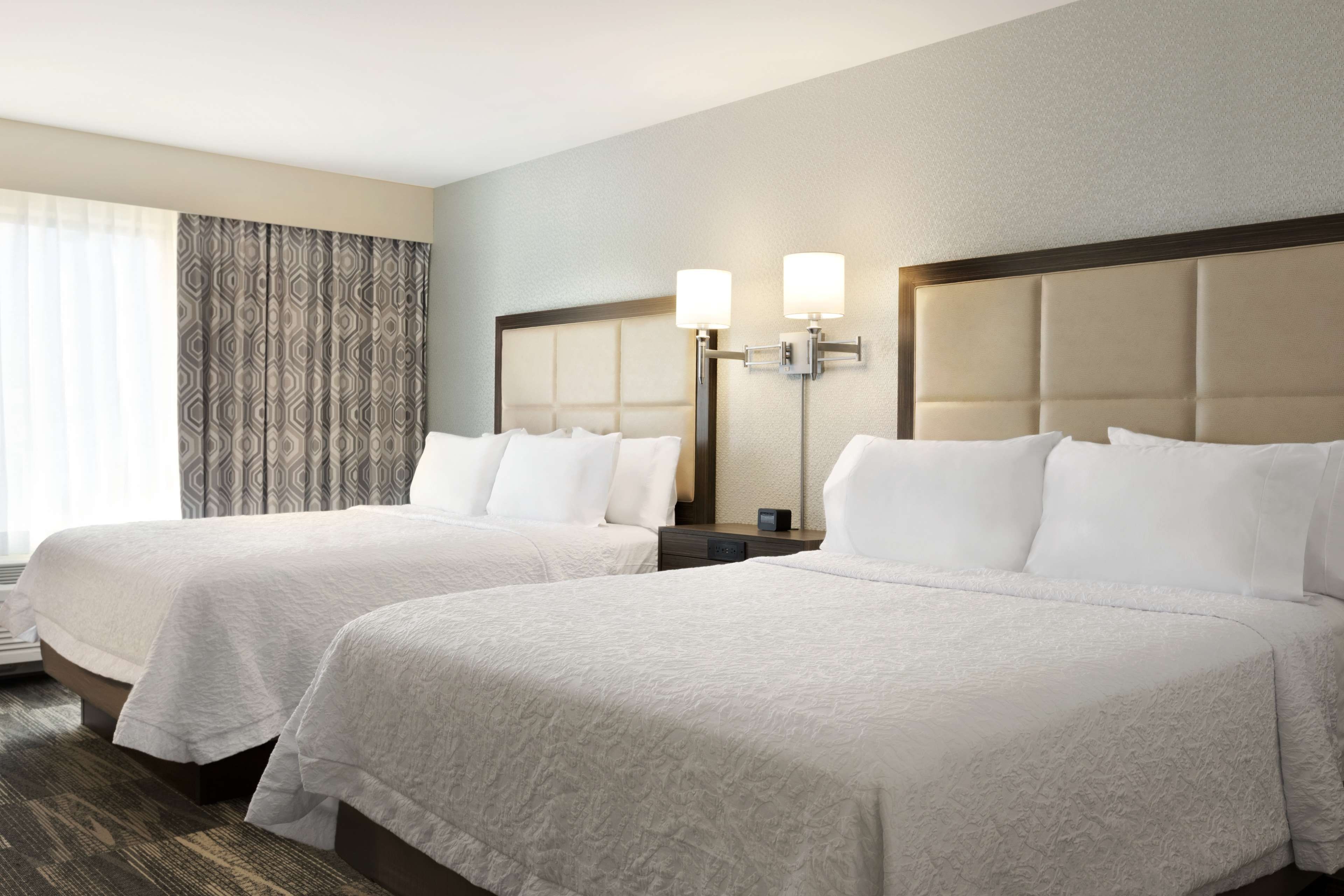 Una habitación de hotel con dos camas, un ventanal con cortinas, una lámpara de pared y suelo alfombrado.