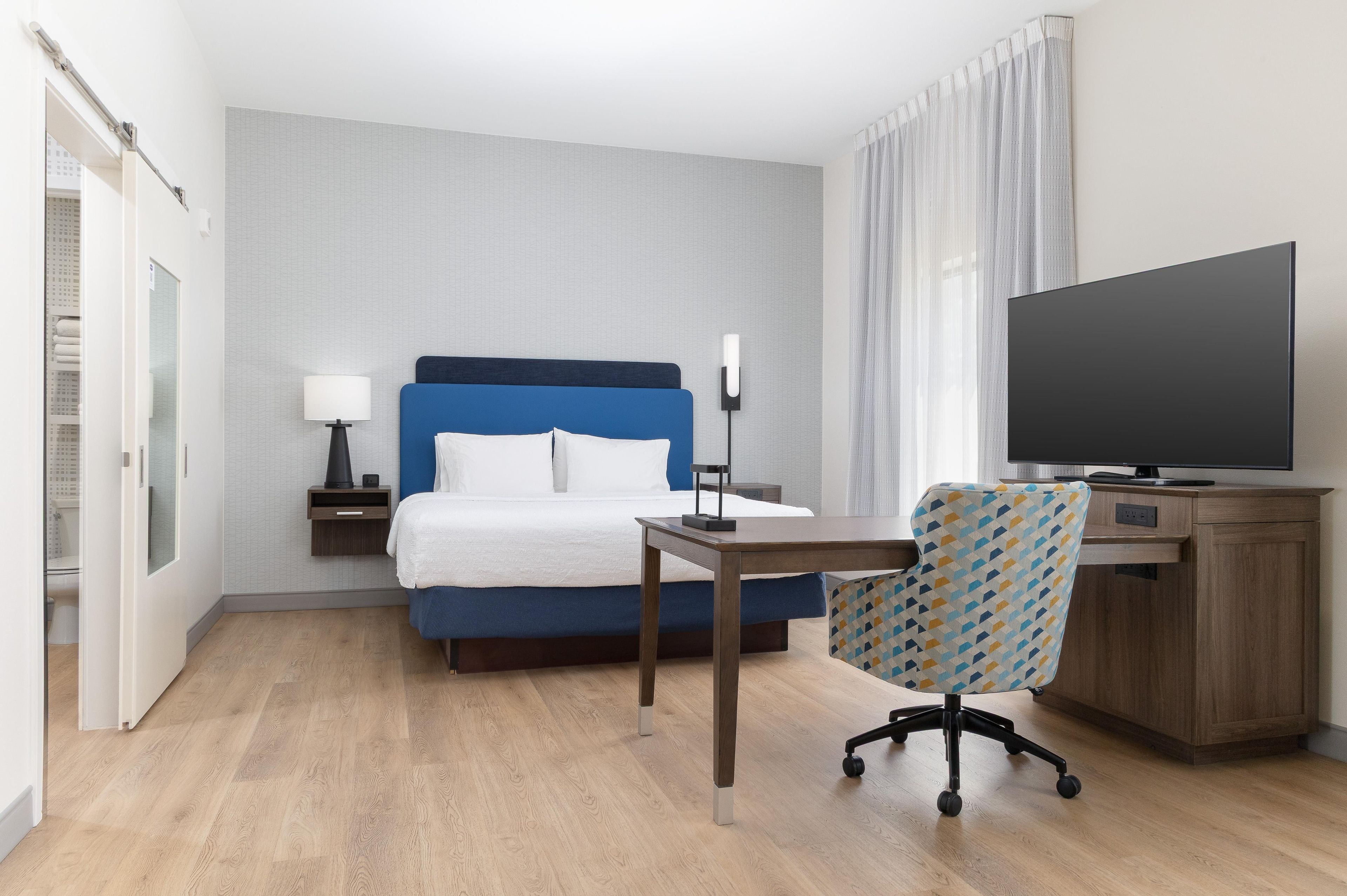 Et hotelværelse med en seng, et skrivebord, en stol, et fjernsyn og et natbord med en lampe.