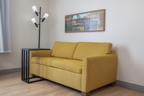 Ein senfgelbes Sofa mit schwarzem Metallgestell, platziert vor einer Wand, über der ein gerahmtes Bild hängt.
