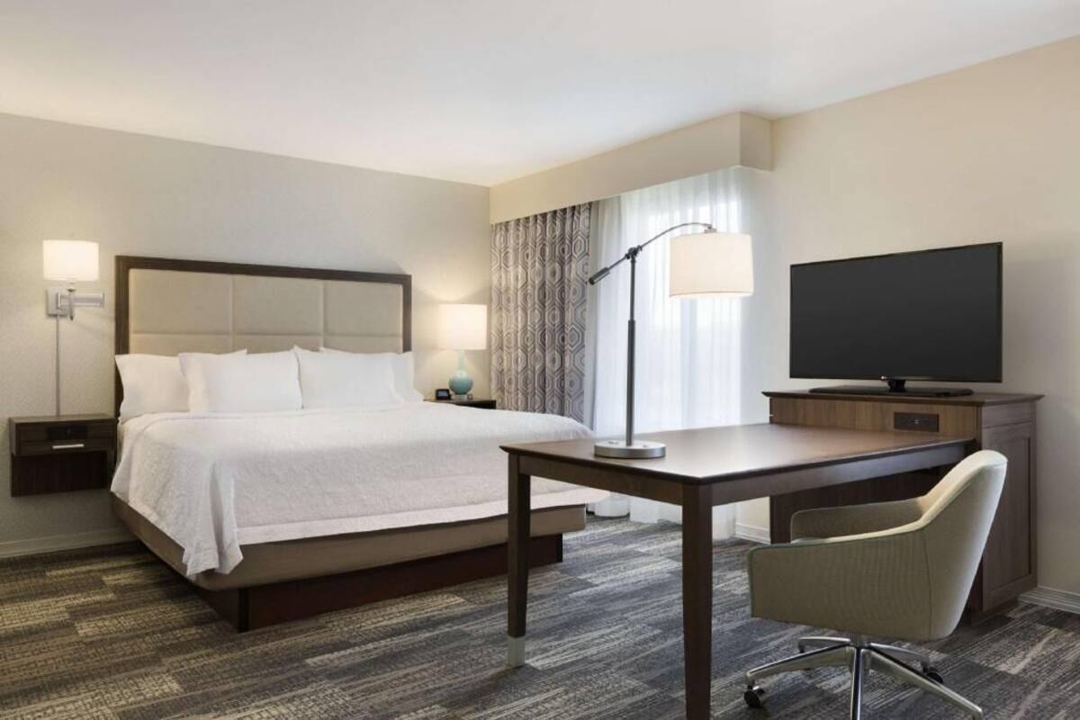 Quarto de hotel com cama, mesas de cabeceira, escrivaninha, cadeira, televisão e luminária.