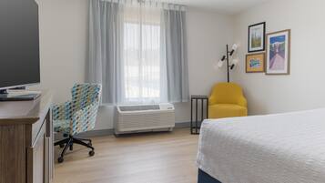 A hotel room with a bed, a desk with a laptop, a chair, an air conditioner, a window with curtains, and framed pictures on the wall.