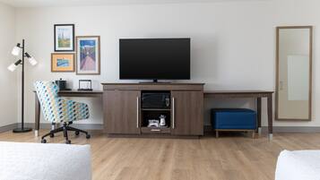 Ein Hotelzimmer mit einem Holztisch, einem Fernseher, einem gemusterten Bürostuhl, einem blauen Hocker und einer Stehlampe.