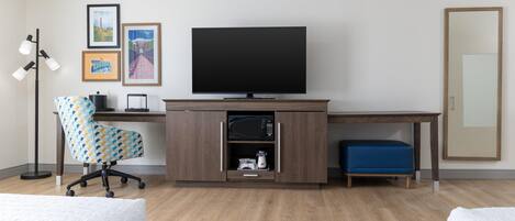A hotel room with a wooden desk, a television, a patterned office chair, a blue ottoman, and a floor lamp.