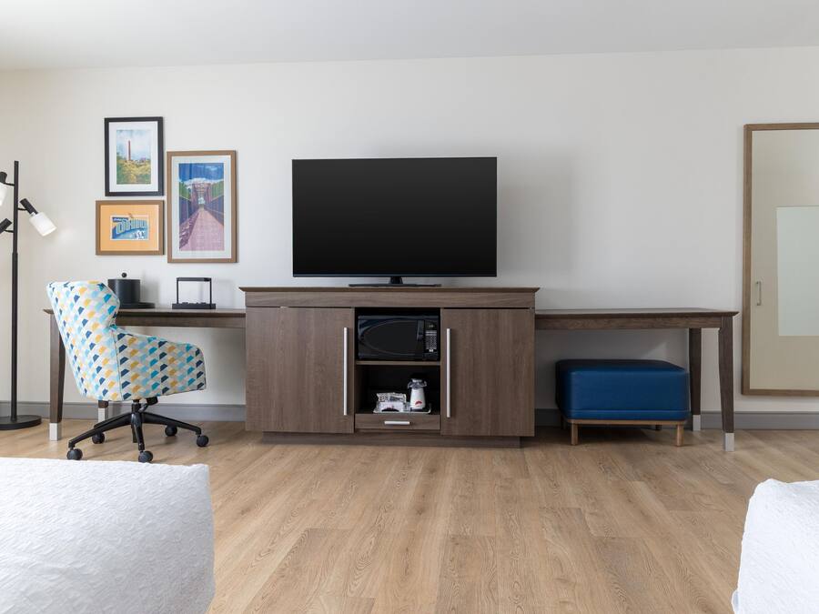 Ein Hotelzimmer mit einem Holztisch, einem Fernseher, einem gemusterten Bürostuhl, einem blauen Hocker und einer Stehlampe.