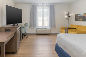 Habitación de hotel con cama, escritorio con televisor de pantalla plana, silla, ventana con cortinas, aire acondicionado, lámpara y un cuadro en la pared.