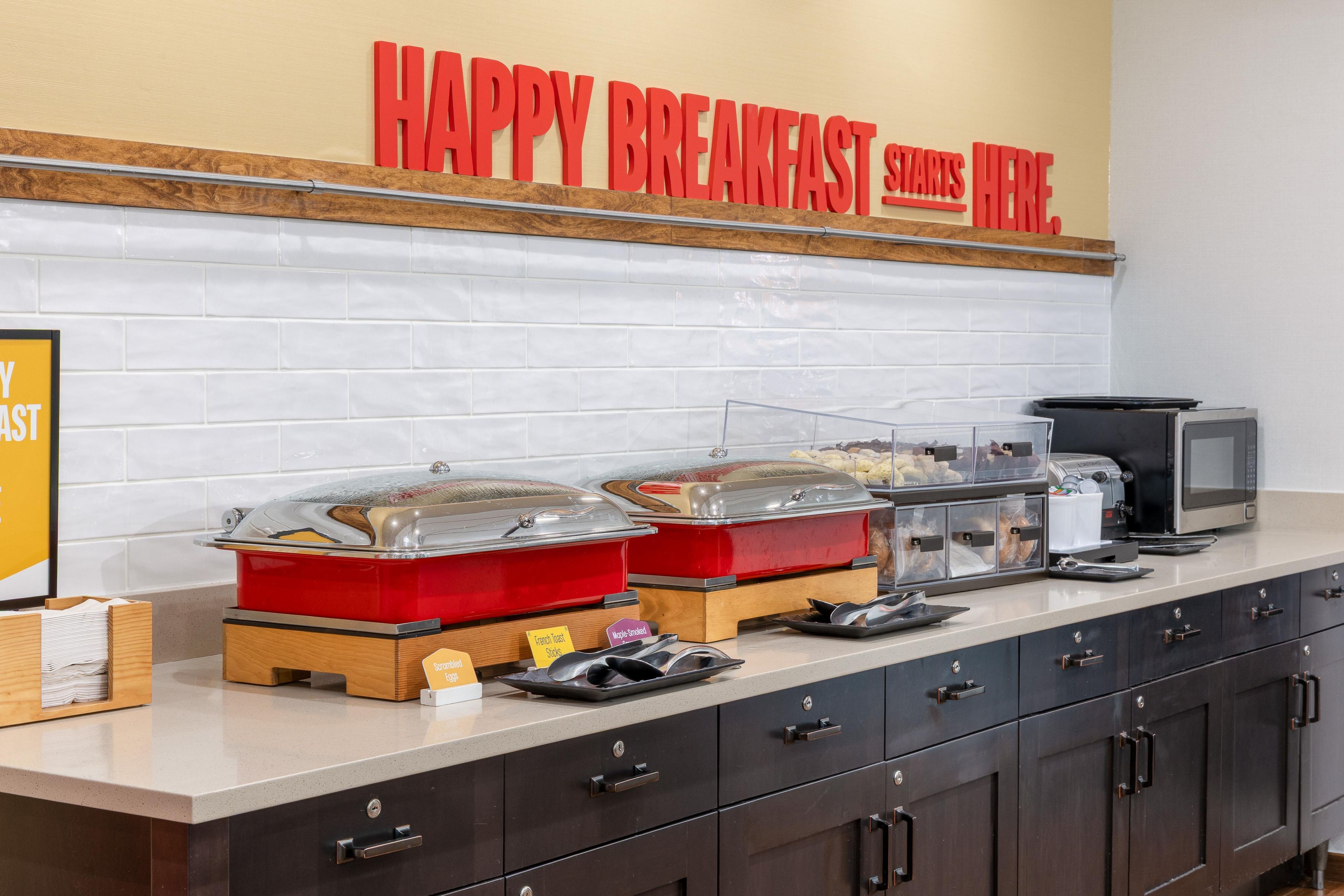 A breakfast buffet setup with labeled containers for French Toast Sticks and Maple-Snicked, a microwave, and a coffee machine.