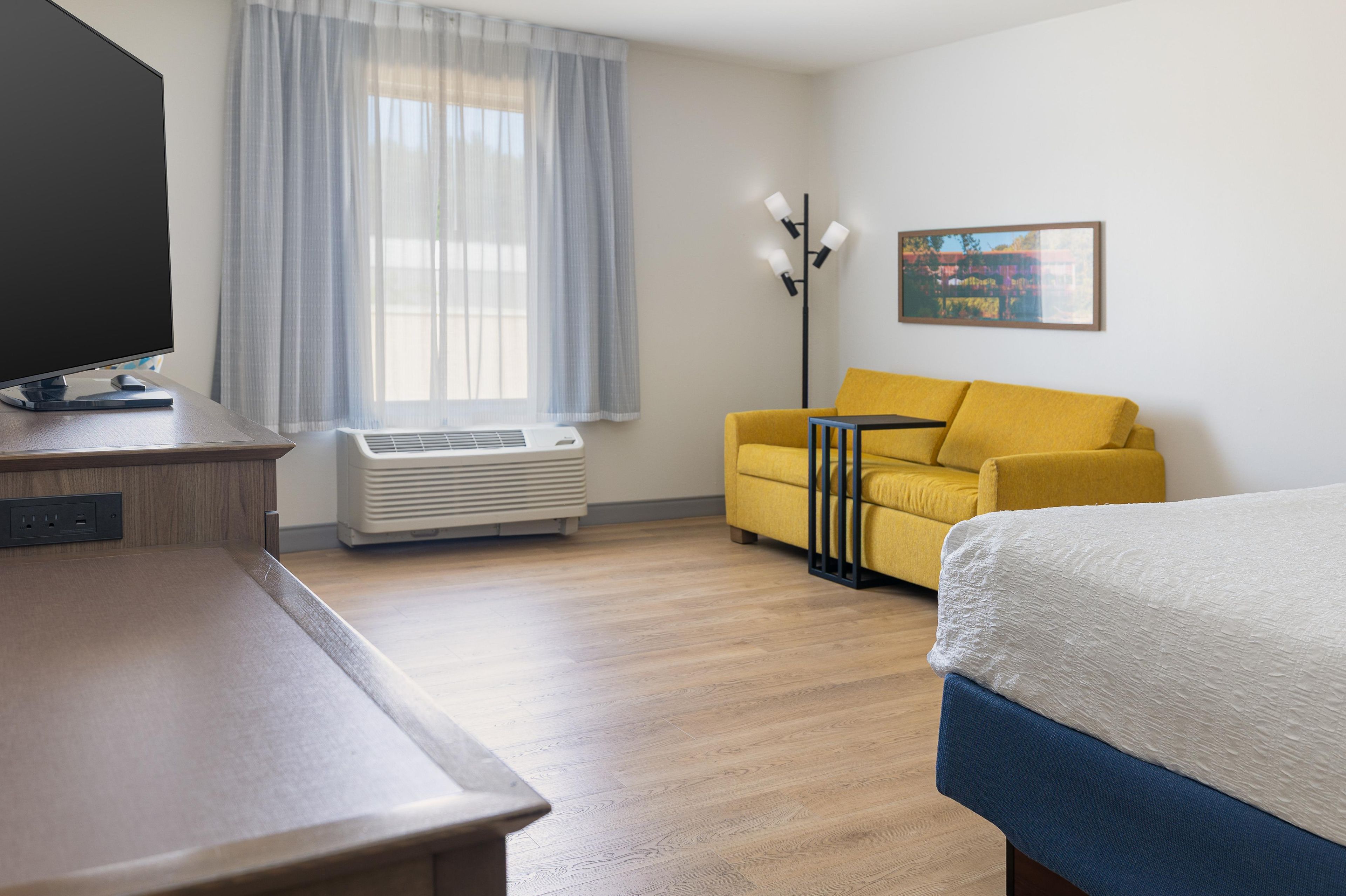 Una habitación de hotel con cama, sofá, escritorio con televisor y ventana con cortinas.