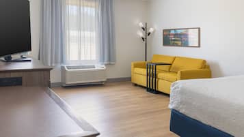 Una habitación de hotel con cama, sofá, escritorio con televisor y ventana con cortinas.