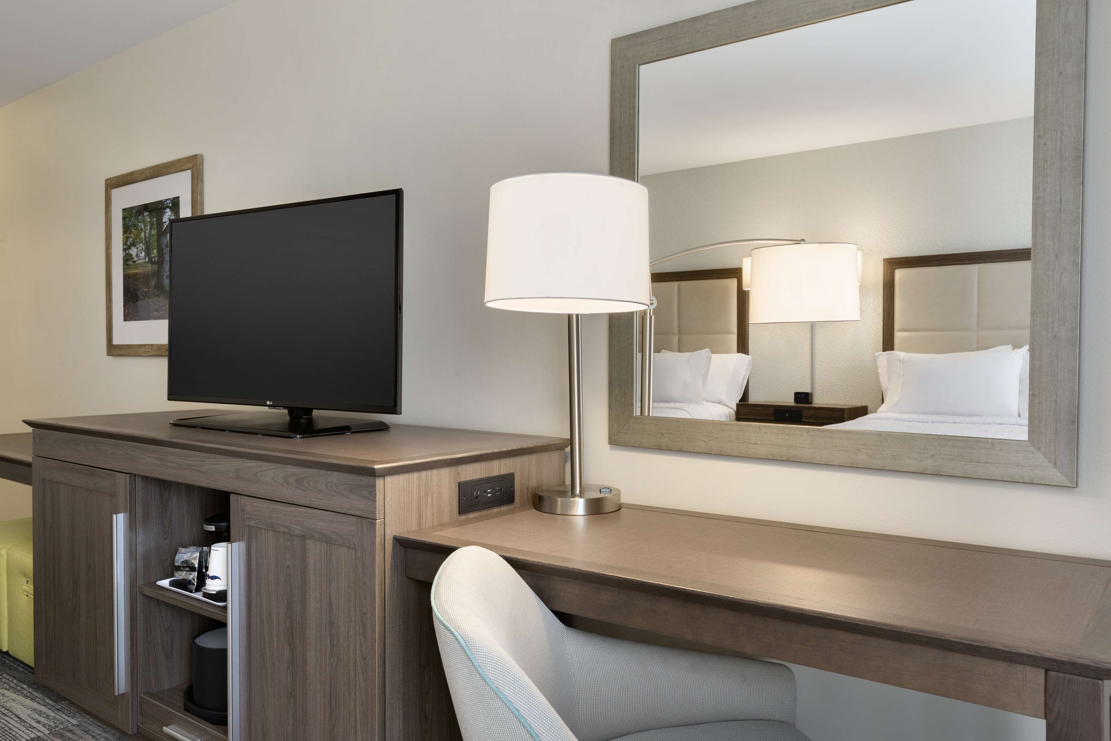 A hotel room with a flat-screen TV, a bedside lamp, a large mirror, a wooden desk, and a chair.