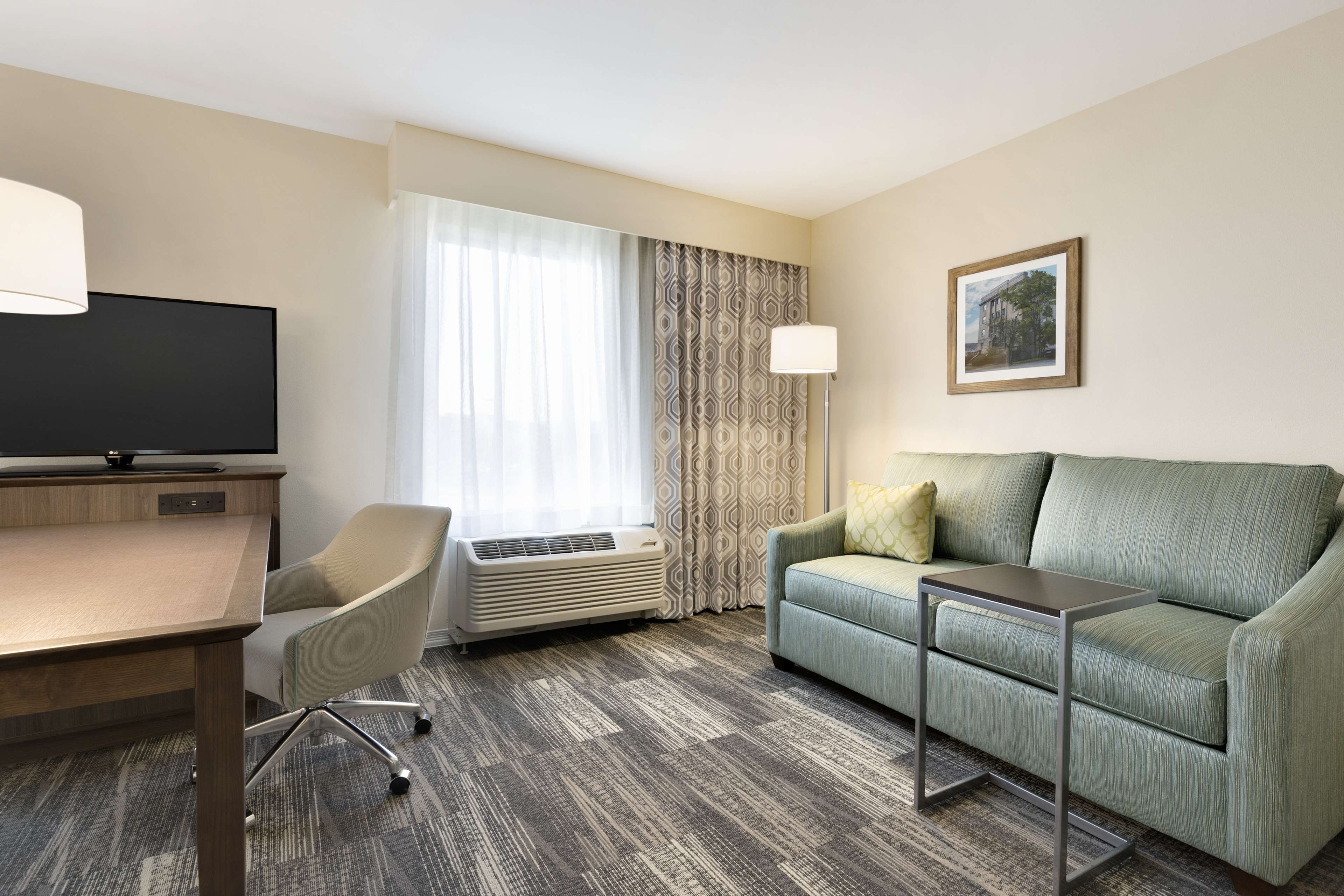 A hotel room with a flat-screen TV, a desk, a chair, a sofa, an air conditioner, a lamp, and a framed picture on the wall.