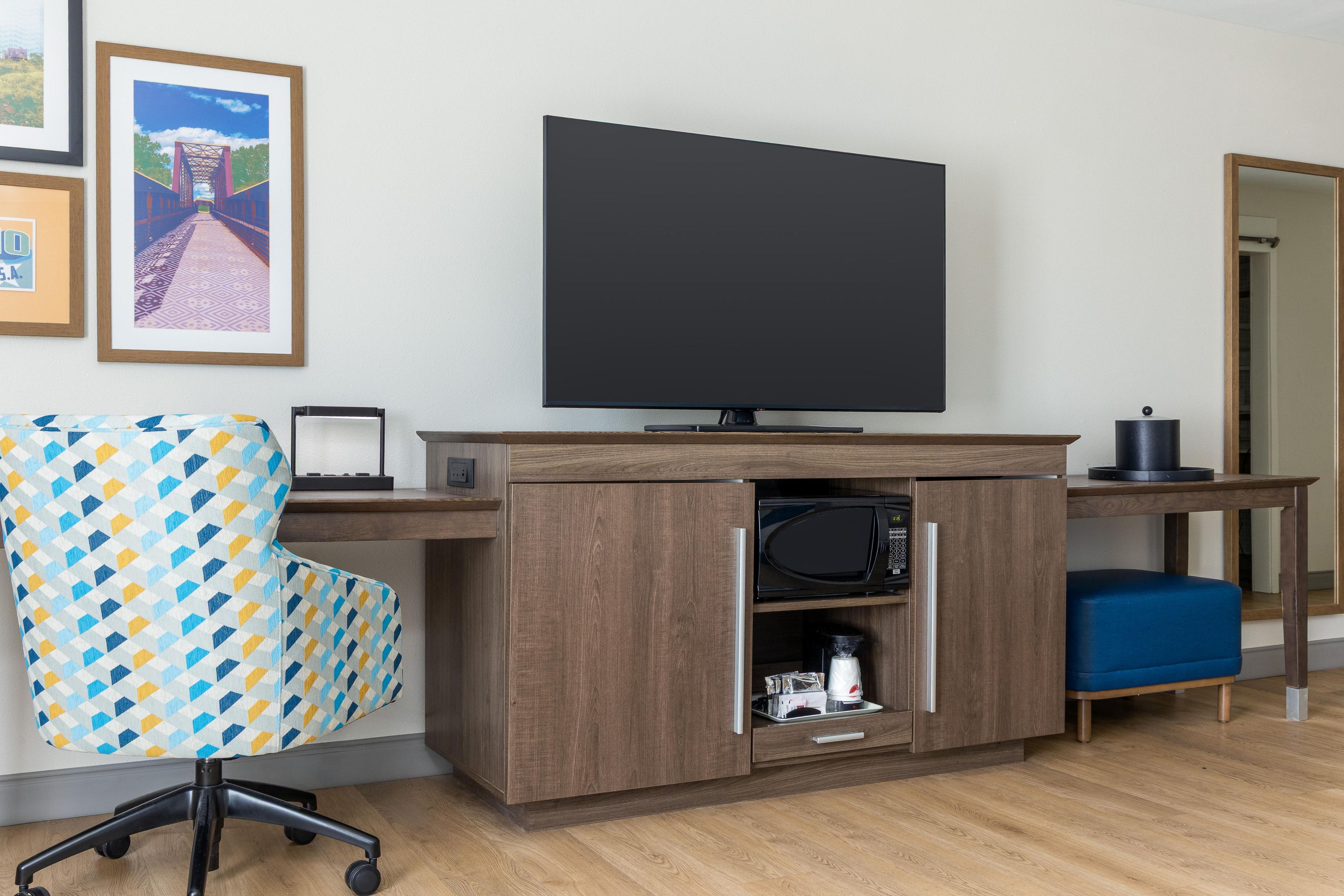 A hotel room with a wooden desk, a flat-screen TV, a patterned armchair, a coffee machine, and a microwave.