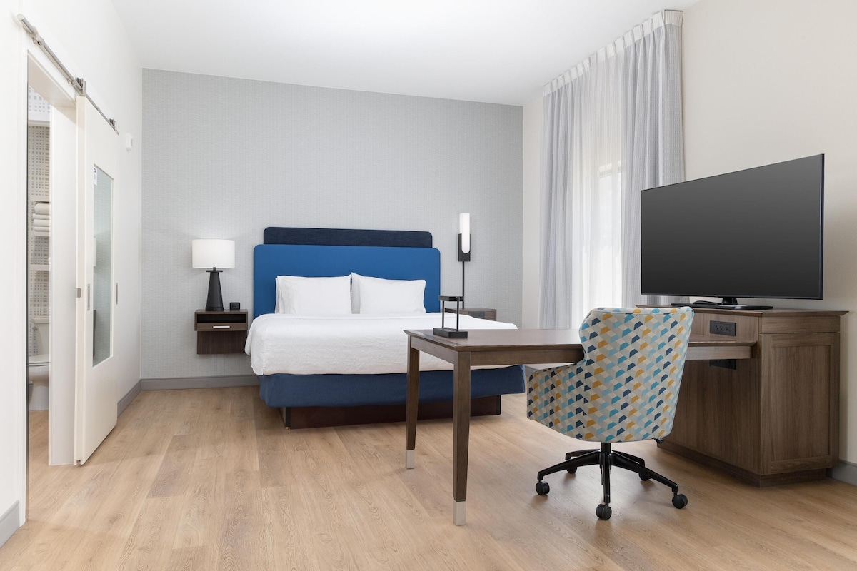 A hotel room with a bed, a desk, a chair, a TV, and a nightstand with a lamp.