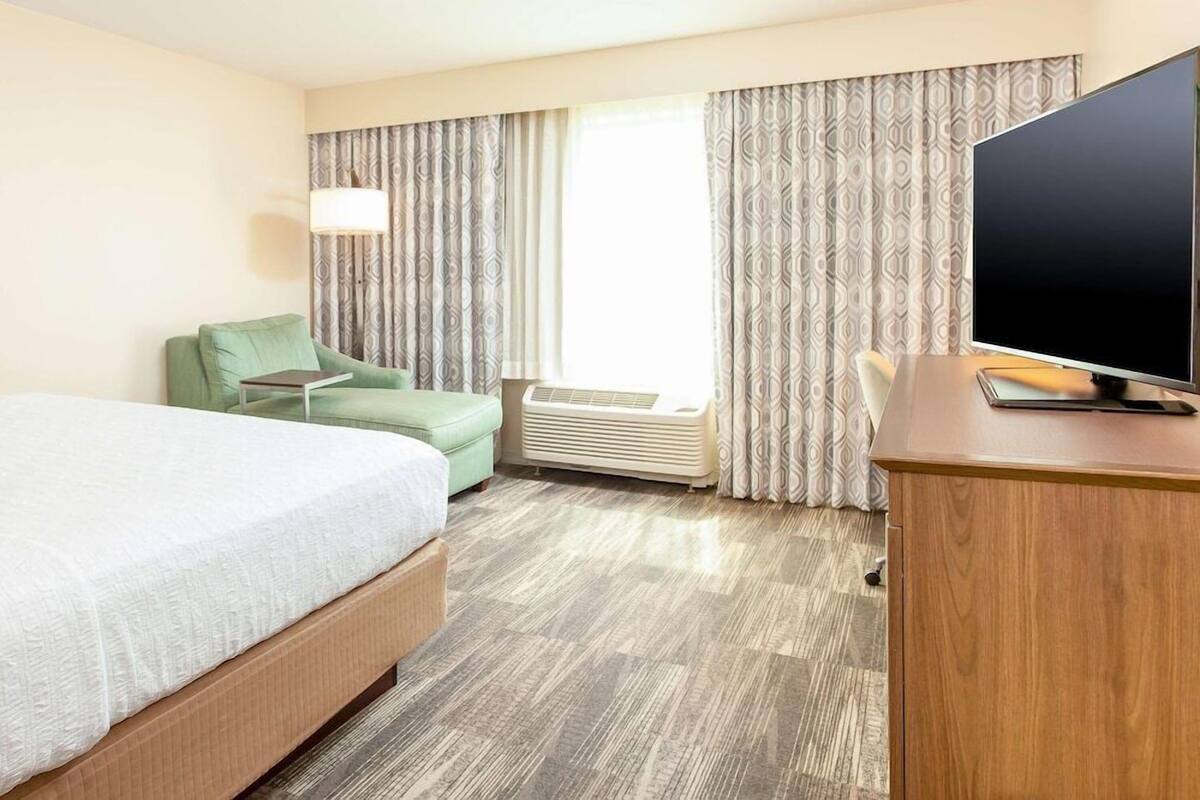 Quarto de hotel com cama, escrivaninha, cadeira, televisão e janela com cortinas.