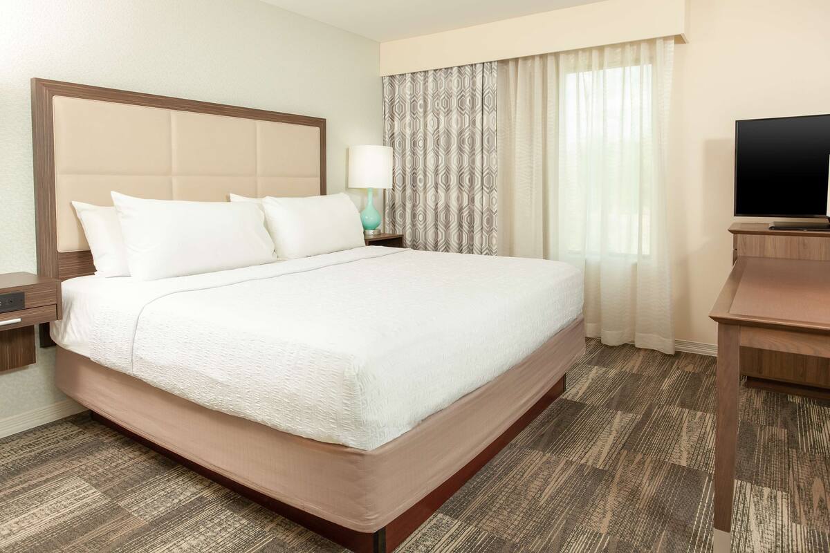 Quarto de hotel com uma cama grande, mesa de cabeceira com abajur, televisão e janela com cortinas.