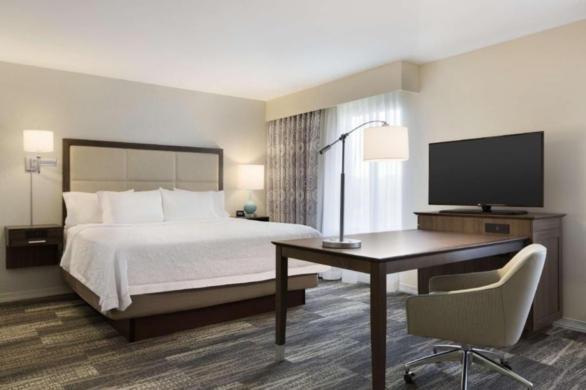 A hotel room with a bed, bedside tables, a desk, a chair, a television, and a lamp.