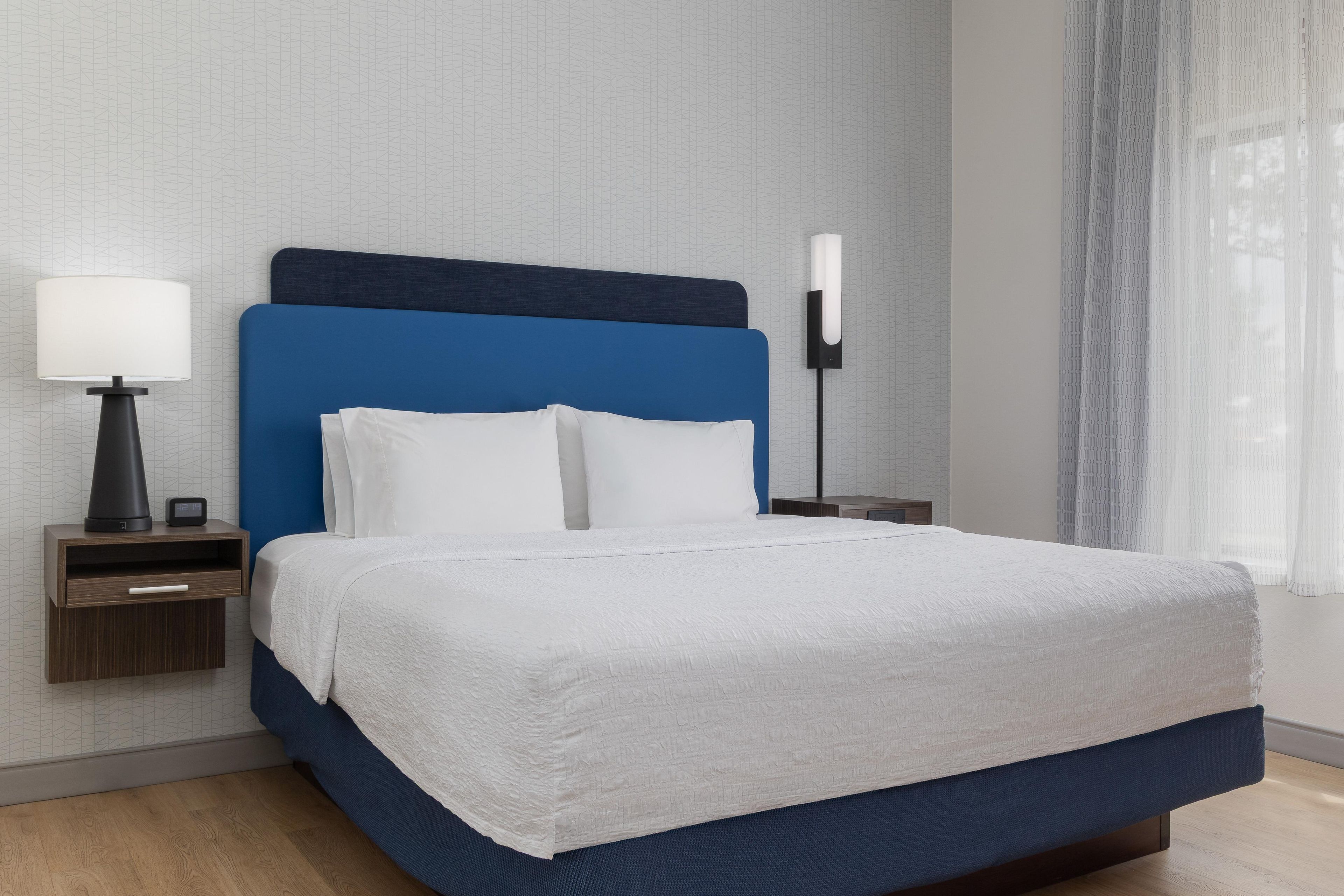 A neatly made bed with a white duvet, a blue headboard, and a bedside lamp on a wooden nightstand.