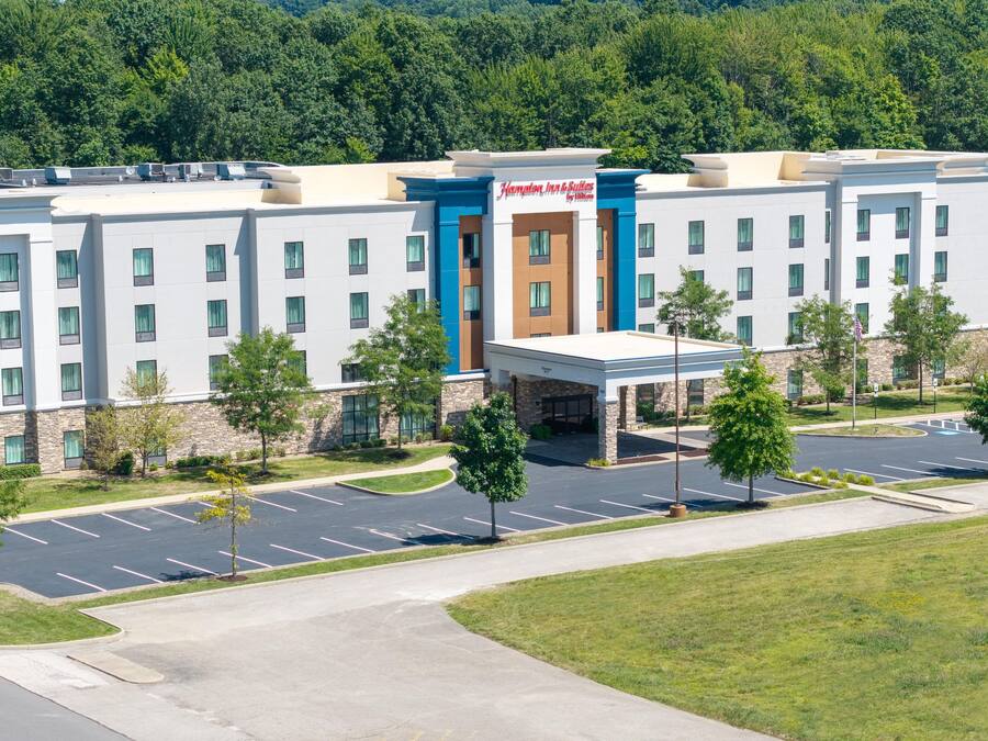 A multi-story hotel with a central entrance, parking lot, and surrounding greenery.