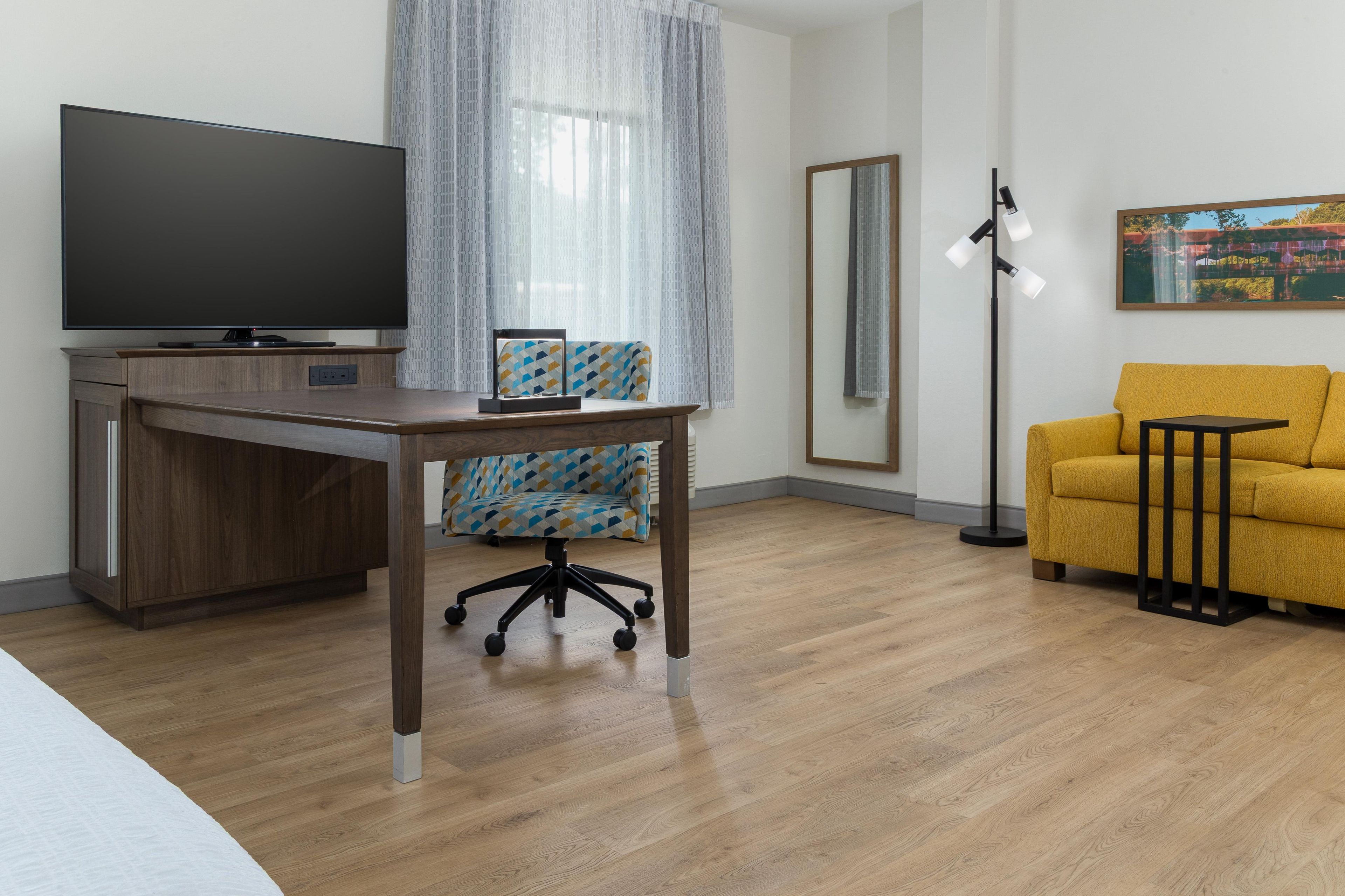 A modern hotel room with a flat-screen TV, a wooden desk, a patterned office chair, a yellow sofa, a floor lamp, and a large mirror.