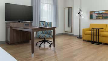 A modern hotel room with a flat-screen TV, a wooden desk, a patterned office chair, a yellow sofa, a floor lamp, and a large mirror.