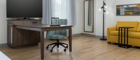 Una habitación de hotel moderna con una televisión de pantalla plana, un escritorio de madera, una silla de oficina estampada, un sofá amarillo, una lámpara de pie y un espejo grande.