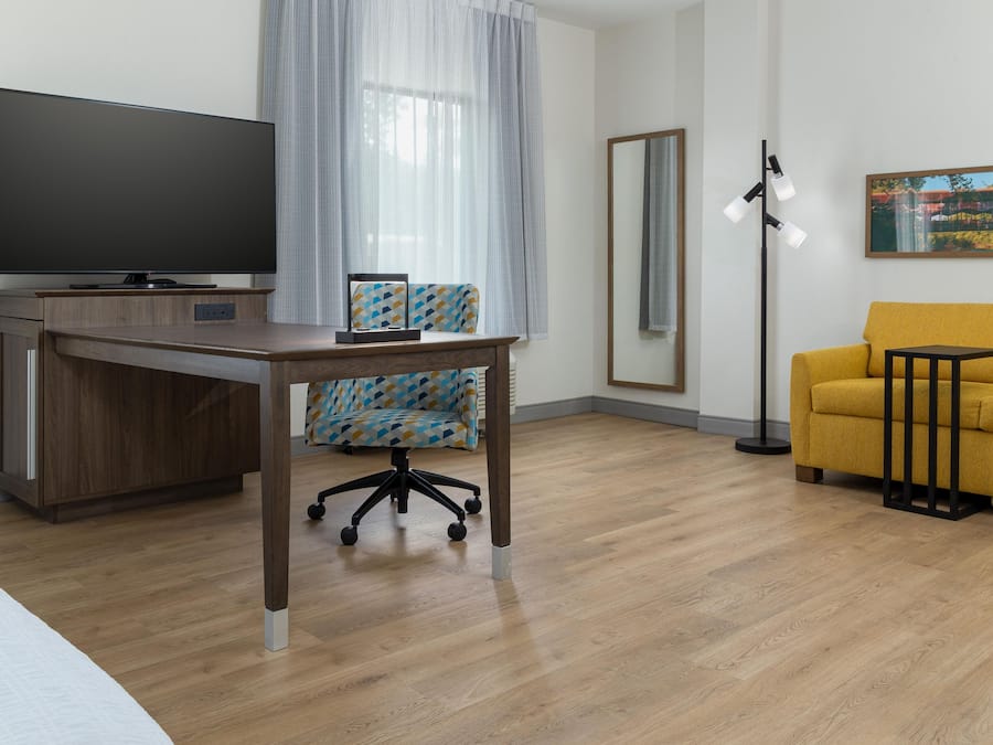 A modern hotel room with a flat-screen TV, a wooden desk, a patterned office chair, a yellow sofa, a floor lamp, and a large mirror.