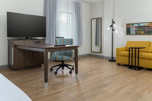 Ett modernt hotellrum med en platt-TV, ett skrivbord i trä, en mönstrad kontorsstol, en gul soffa, en golvlampa och en stor spegel.