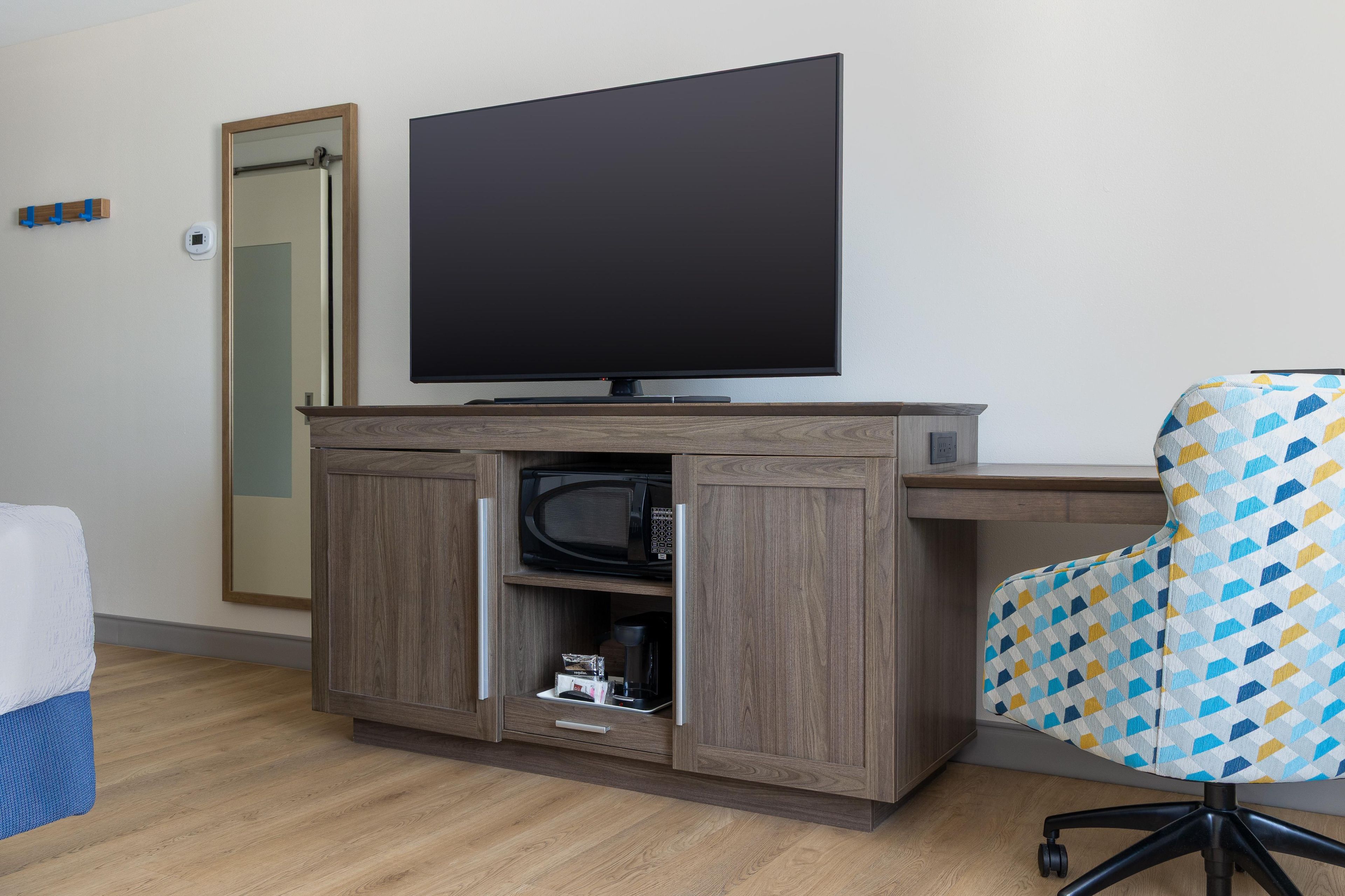 Une chambre d’hôtel équipée d’un téléviseur à écran plat fixé à un meuble en bois, d’un four à micro-ondes, d’un petit tiroir et d’un fauteuil à motifs.