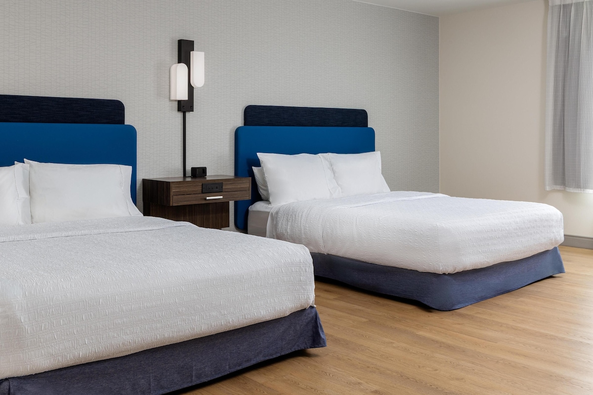 A hotel room with two beds, a nightstand, a lamp, and a window with curtains.