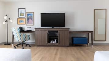 A hotel room with a wooden desk, a television, a patterned office chair, a blue ottoman, and a floor lamp.