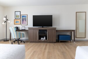 Habitación de hotel con un escritorio de madera, una televisión, una silla de oficina estampada, un otoman azul y una lámpara de pie.