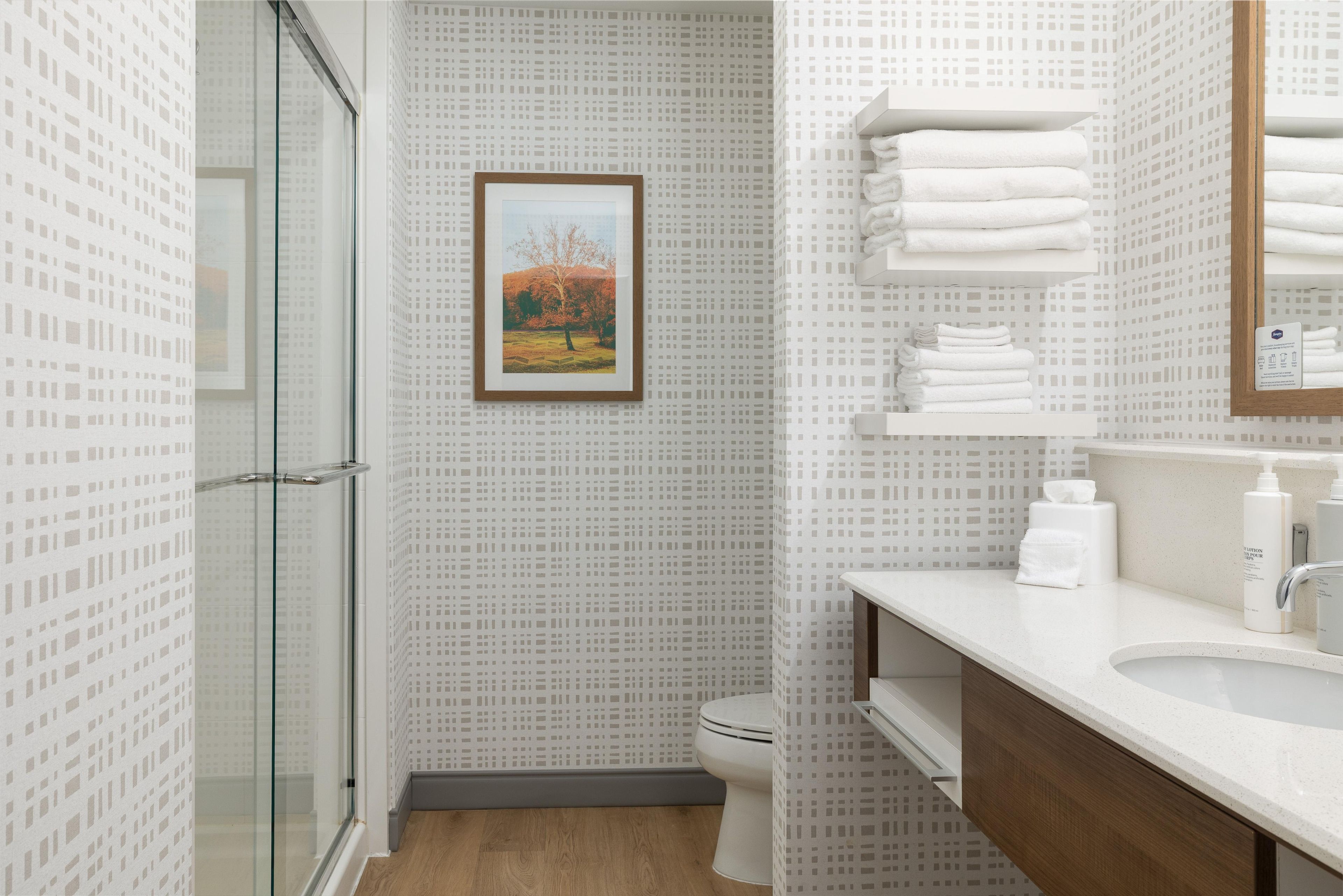 A bathroom with a glass shower enclosure, a toilet, a sink with toiletries, and a framed picture on the wall.
