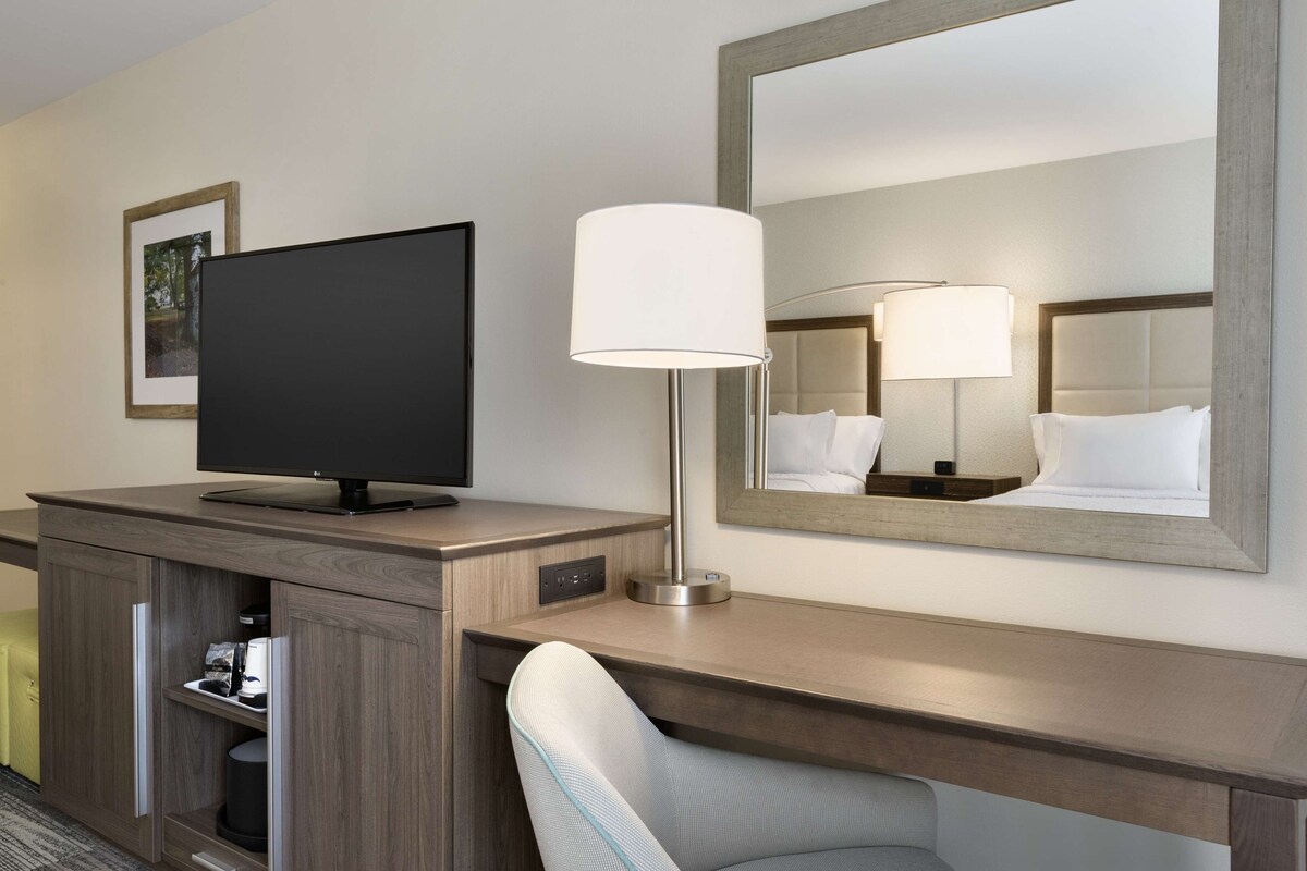 A hotel room with a flat-screen TV, a bedside lamp, a large mirror, a wooden desk, and a chair.