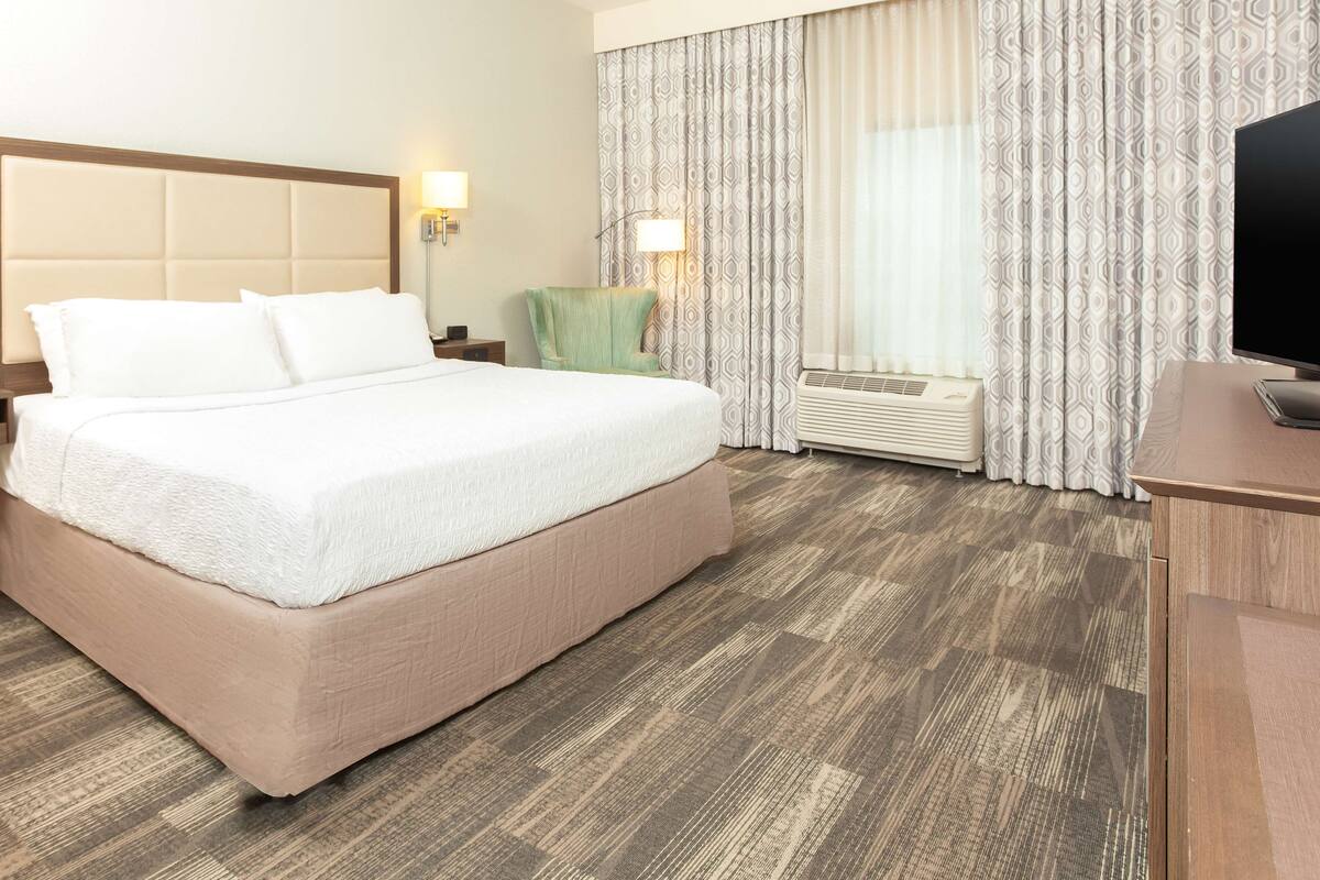 Quarto de hotel com uma cama grande, mesa de cabeceira, luminária, cadeira, televisão e janela com cortinas.