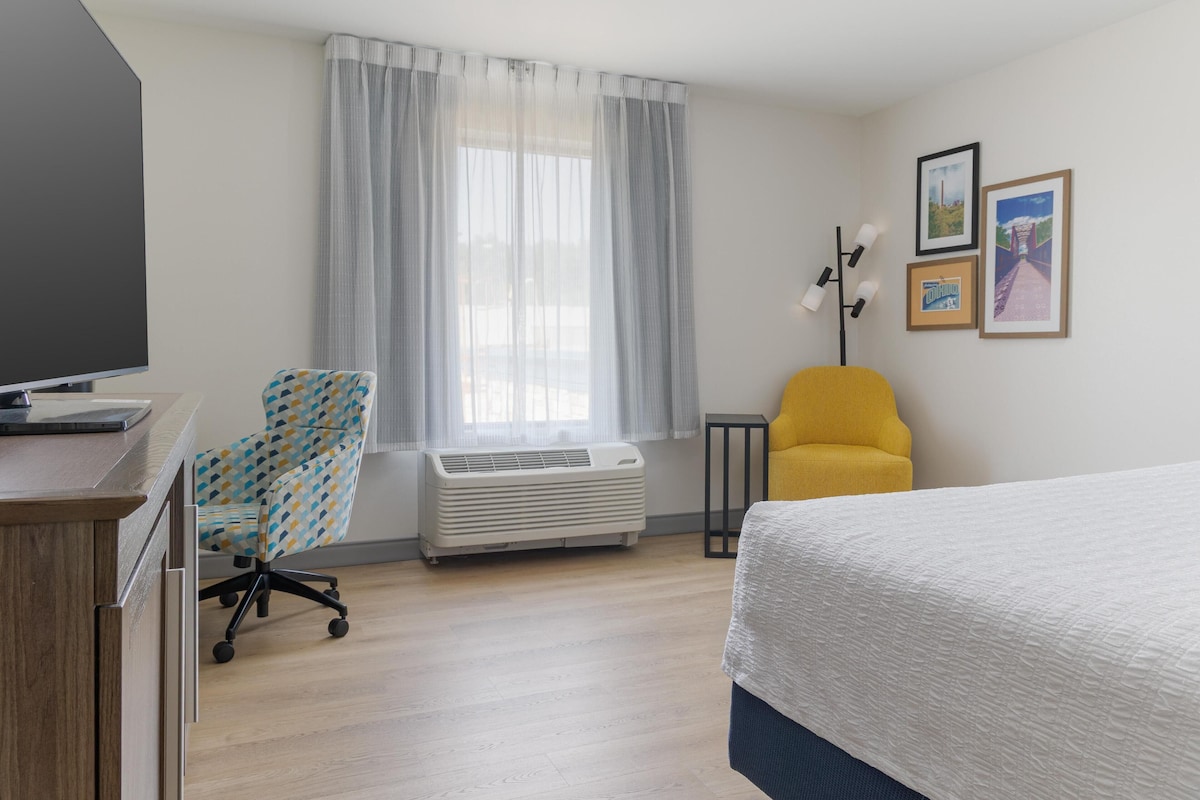 A hotel room with a bed, a desk with a laptop, a chair, an air conditioner, a window with curtains, and framed pictures on the wall.
