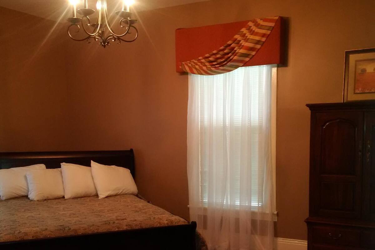 Un dormitorio con cama, un candelabro, una ventana con cortinas translúcidas y un armario de madera.