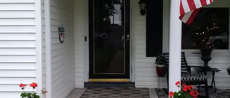 Uma casa branca com o número 105 na porta da frente, uma porta preta, um tapete de boas-vindas e dois vasos de plantas com flores vermelhas na varanda.