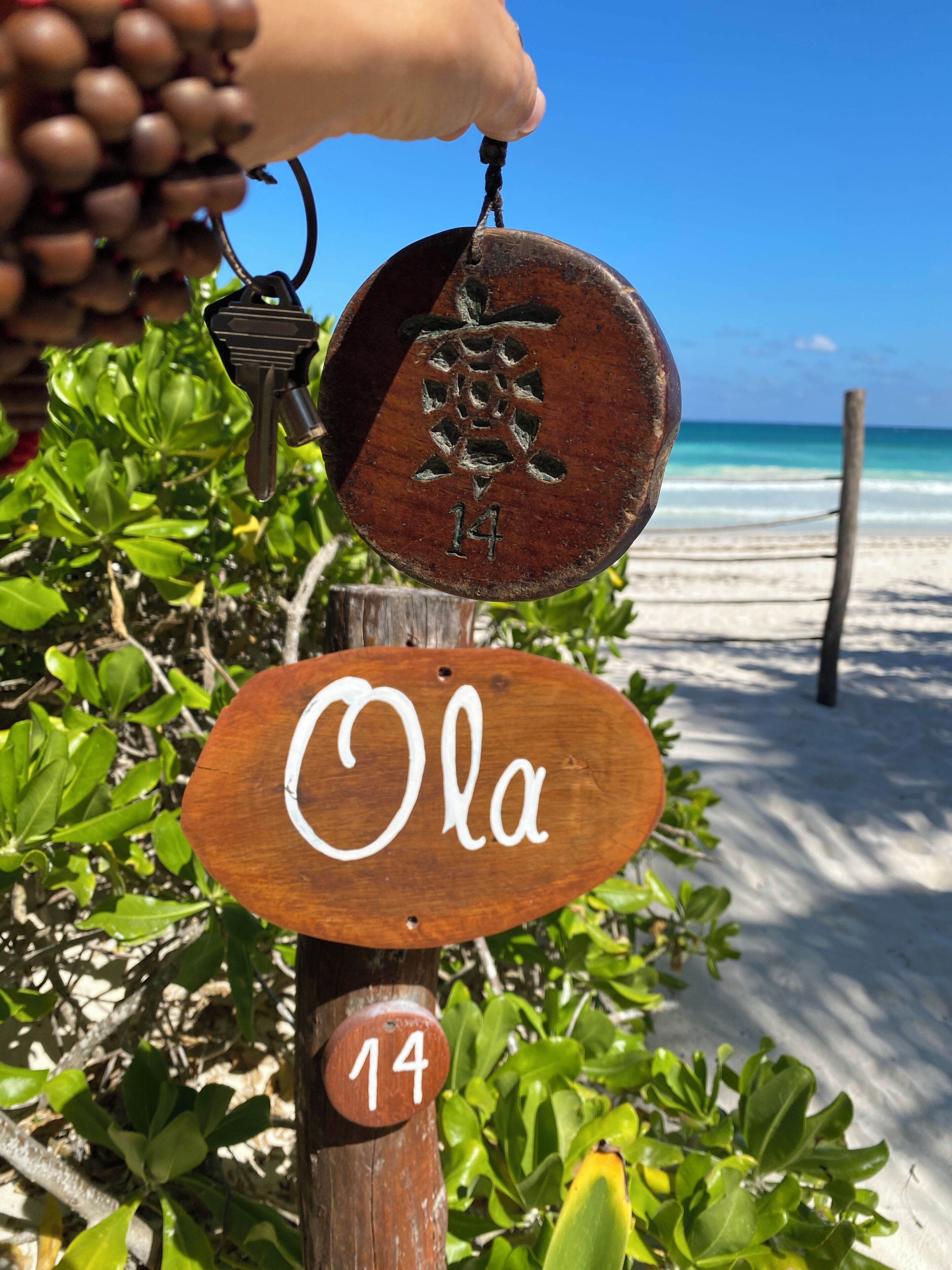 A wooden sign with the name "Ola" and the number "14" on a post, with a beach and ocean in the background.