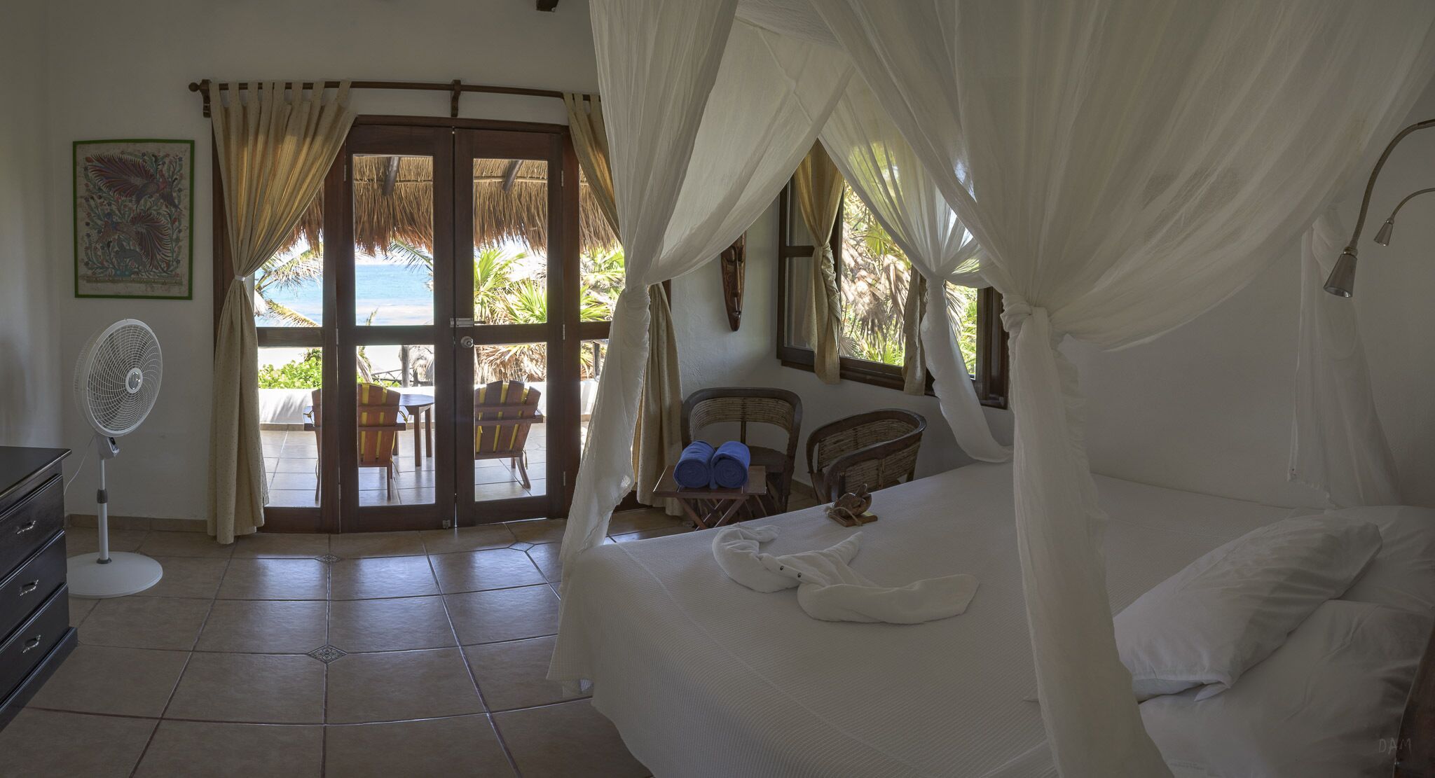 Una habitación con cama, ventilador y vista a la playa a través de un ventanal.