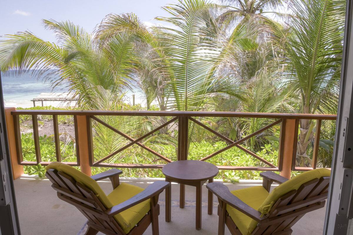 A balcony with wooden chairs and a table, overlooking a beach and palm trees.
