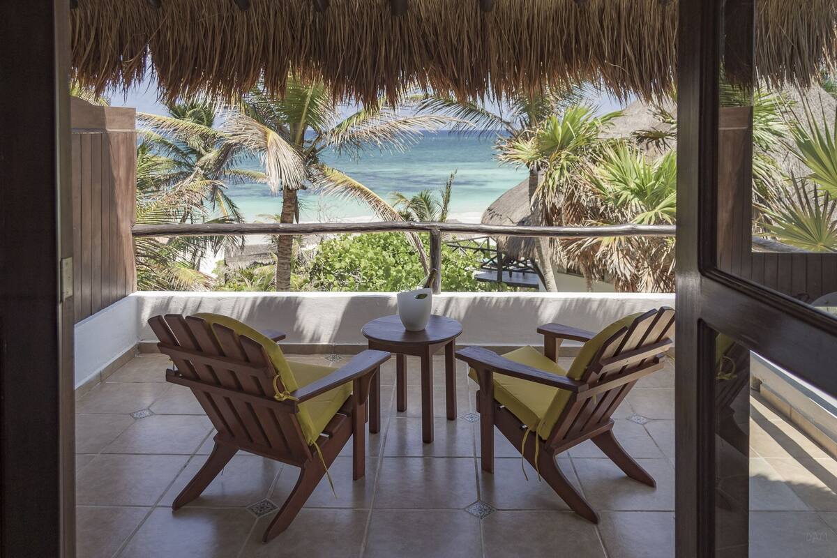 A balcony with wooden chairs and a small table, overlooking a beach with palm trees and the ocean.