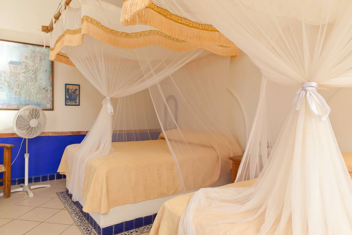 A room with two beds, each with a canopy bedspread and a mosquito net. There is a standing fan and a blue tiled floor.