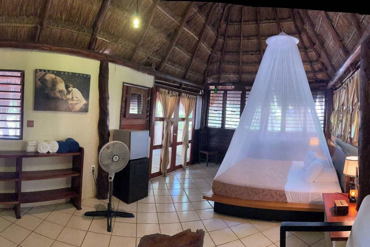 A room with a thatched roof, a bed with a mosquito net, a fan, and a small table with a lamp.