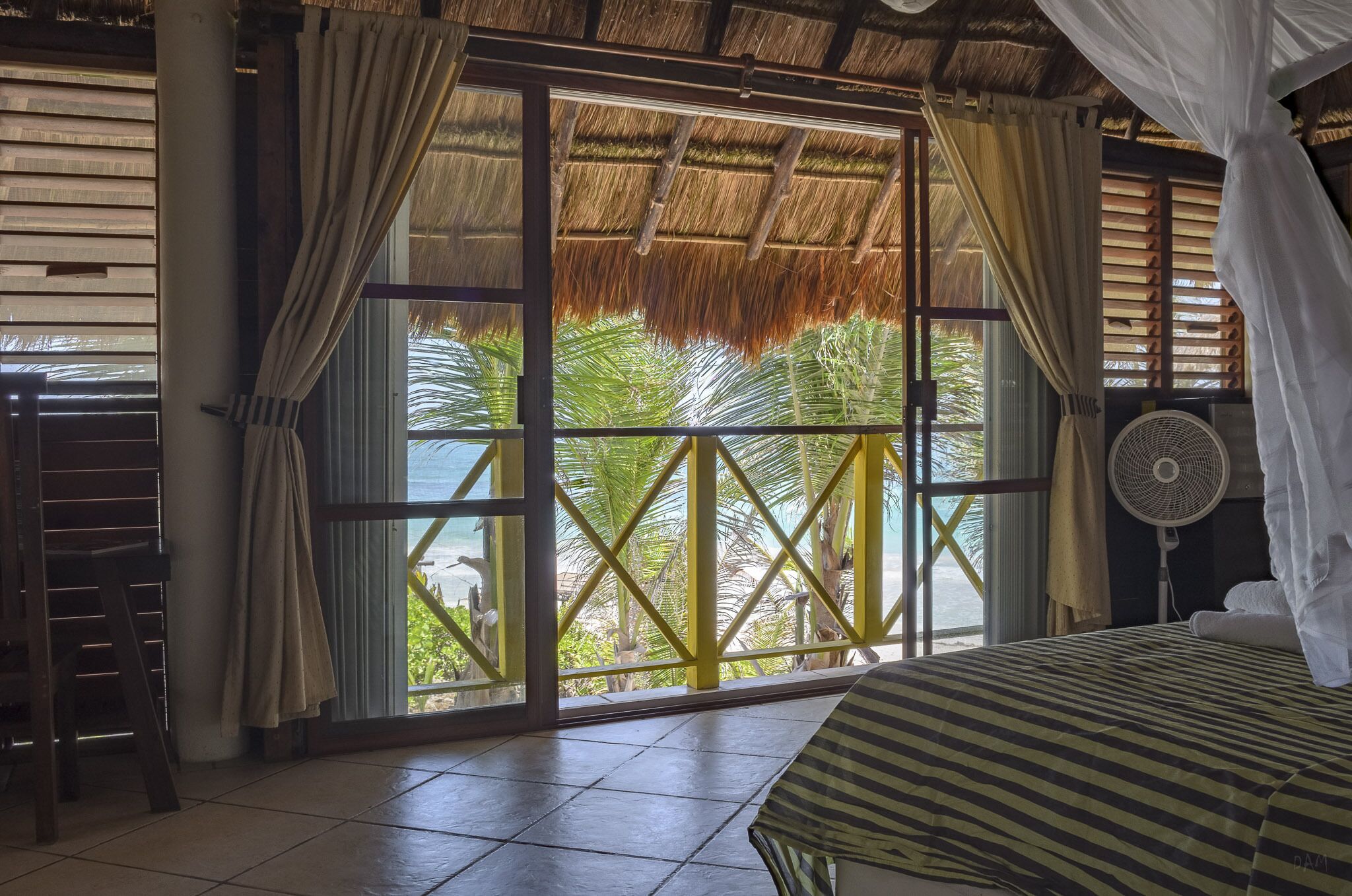 Une chambre avec un lit, un ventilateur et une vue sur les palmiers, visible à travers une grande fenêtre.