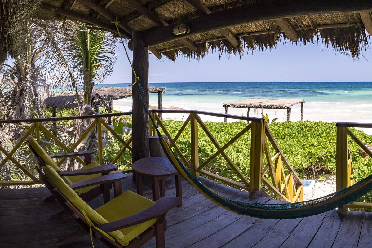 A wooden deck with a hammock, chairs, and tables overlooking a beach with clear blue water and a thatched roof structure.